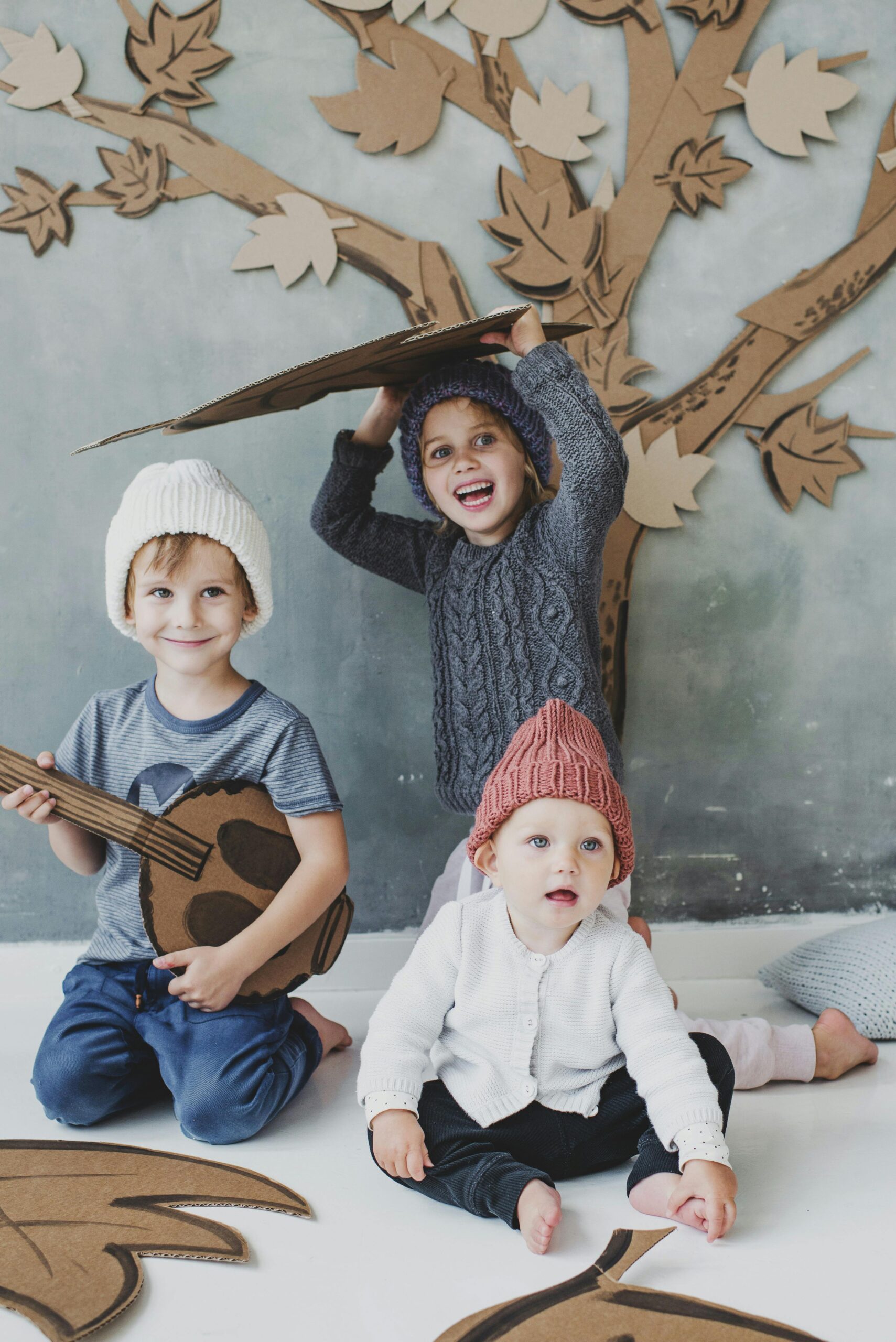 Three kids enjoying playtime with cardboard cutouts indoors, wearing knitted hats.