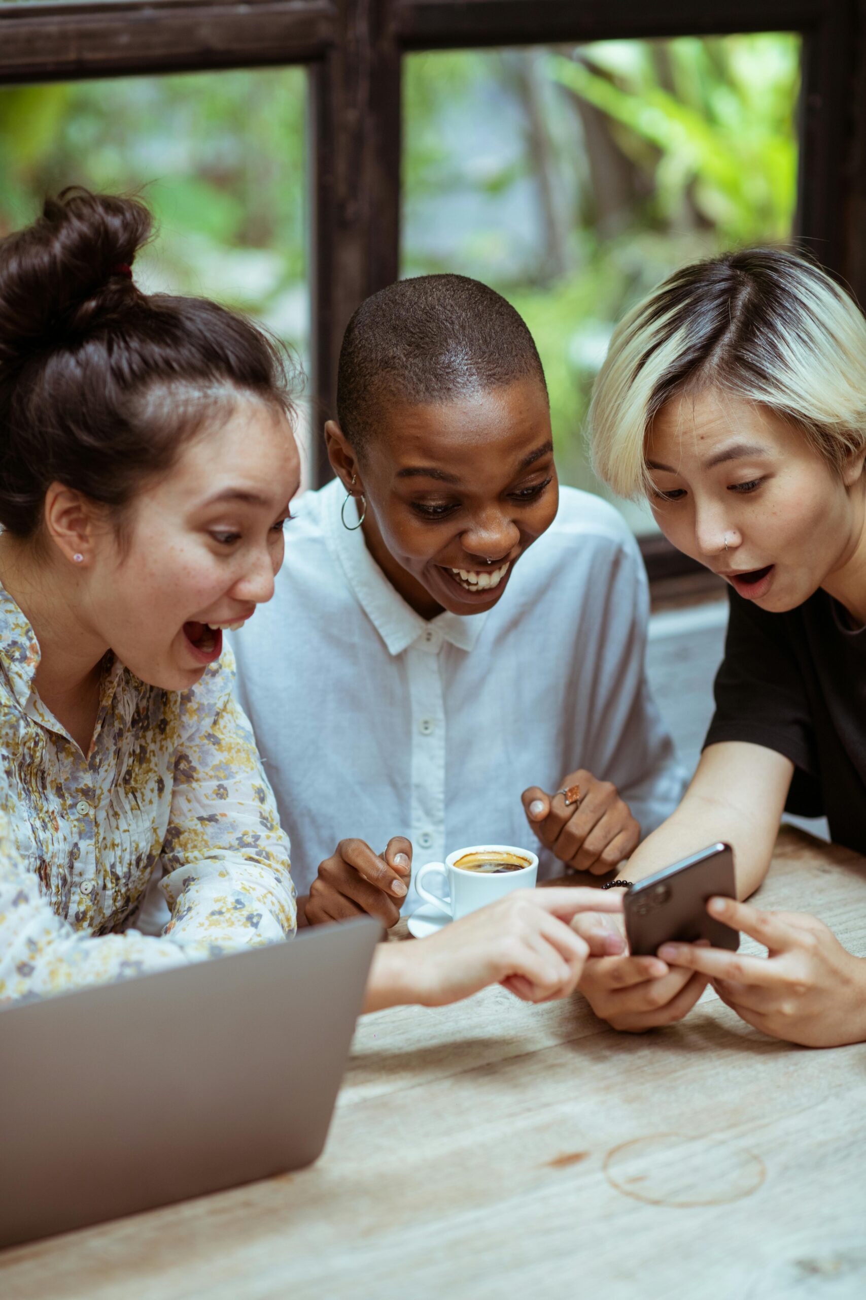 Amazed surprized multiethnic female friends in casual clothes with cup of coffee browsing smartphone while sitting at wooden table with laptop against window