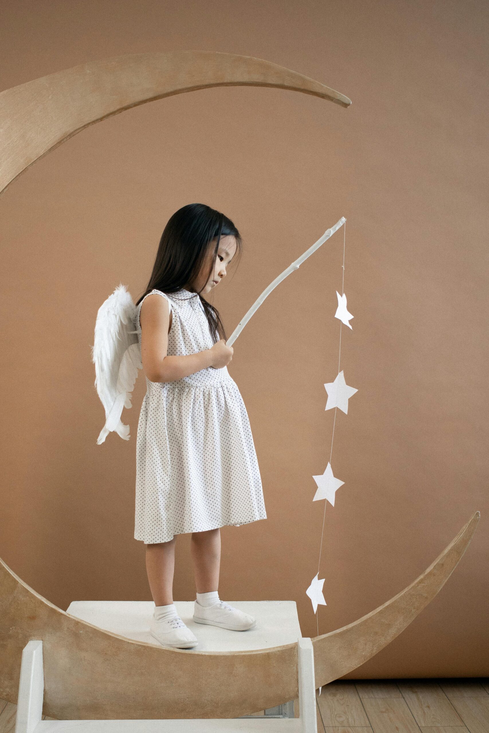 A young girl dressed as an angel, standing on a crescent moon with hanging stars.