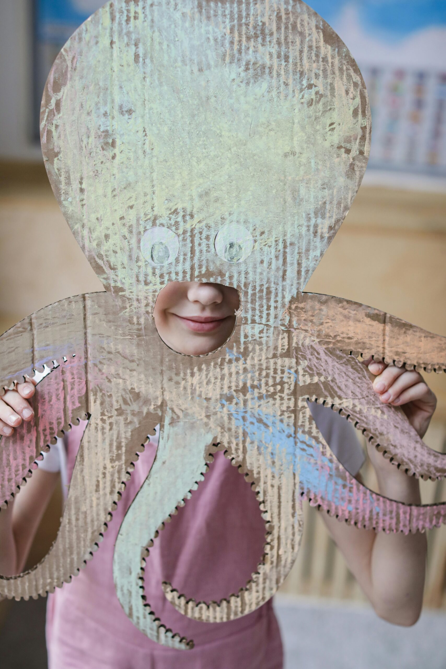 Young child holding a handmade cardboard octopus mask indoors, capturing joyful creativity.